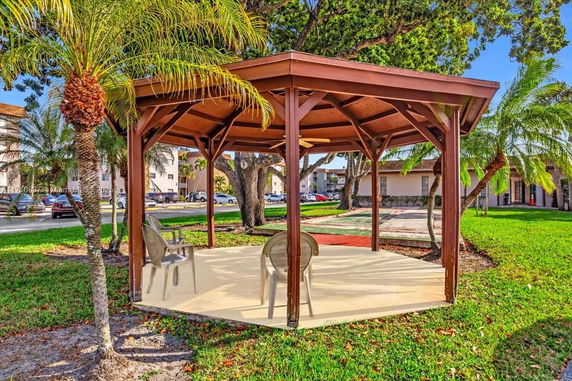Outdoor view of a wooden hexagonal gazebo with chairs on a concrete slab in a green area.