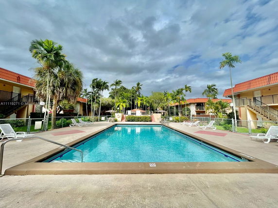 Outdoor swimming pool surrounded by apartment buildings with palm trees and lounge chairs.