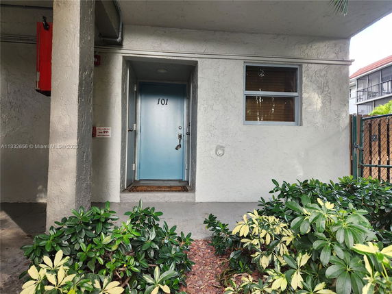 Front entrance view of a residential unit with a blue door and window, surrounded by greenery.