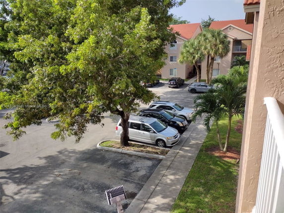 View of an outdoor parking area with several cars parked. Residential buildings and trees in the background.