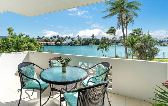 View of a waterfront area from a balcony, featuring outdoor seating and lush greenery.