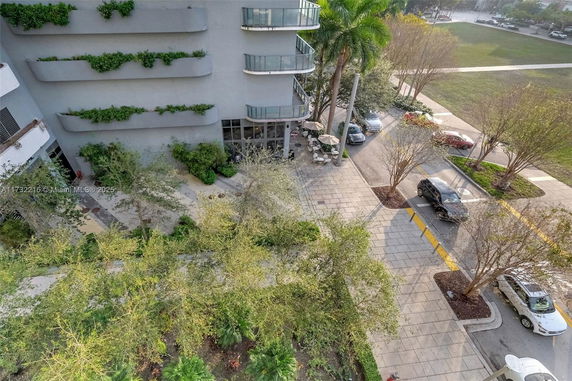 Aerial view of a building with balconies and green planters, overlooking a street with parked cars and surrounding trees.