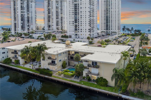 Aerial view of a waterfront residential building with surrounding high-rise structures and a nearby coastline.