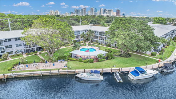 A view of a residential area with apartment buildings surrounding a circular swimming pool, positioned near a waterfront lined with boats.