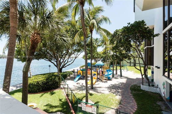 Outdoor playground with slides and play structures near a water body and surrounded by palm trees.