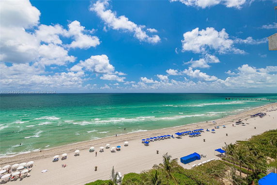 Panoramic view of a beach with turquoise water, sand, umbrellas, and a pier in the distance.