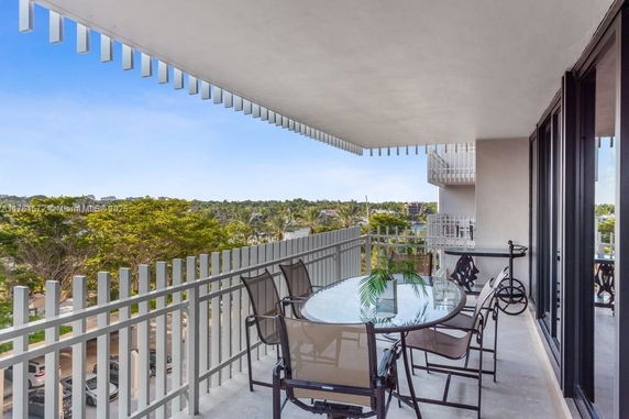 A balcony with a glass table and chairs, offering a view of the surrounding trees and buildings.