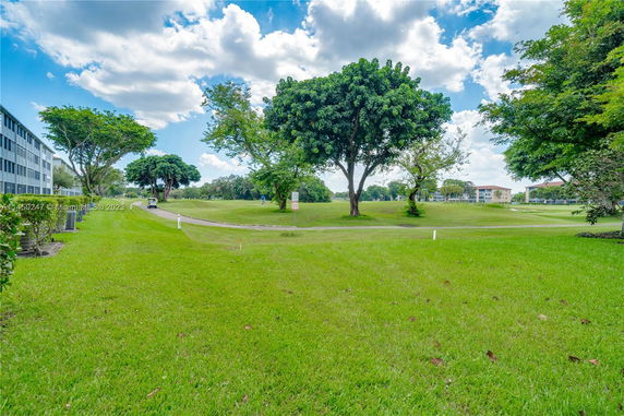 View of a grassy landscape with trees and a few buildings in the distance under a partly cloudy sky.