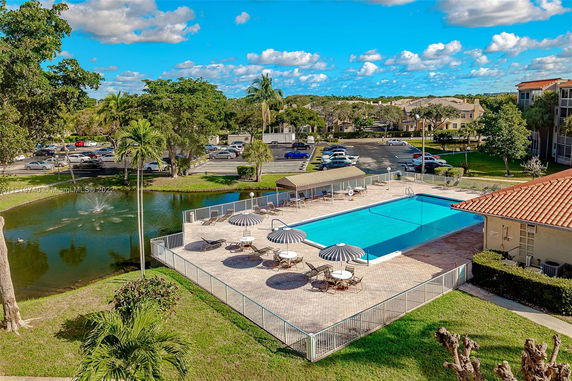 Outdoor swimming pool area with sun umbrellas, tables, and chairs set nearby, adjacent to a parking lot and pond.