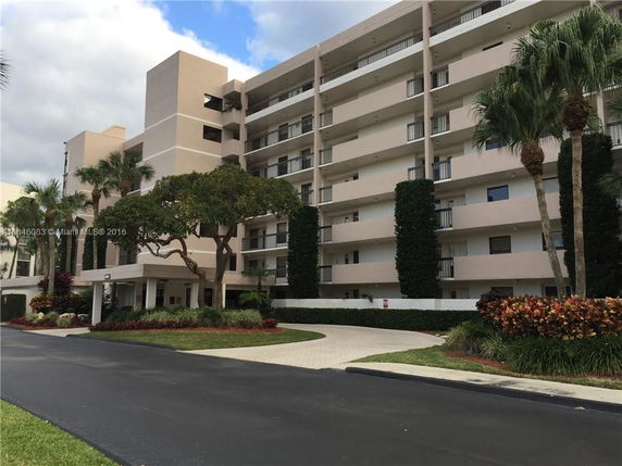 Front view of a multi-story residential building with balconies and surrounded by trees and landscaping.