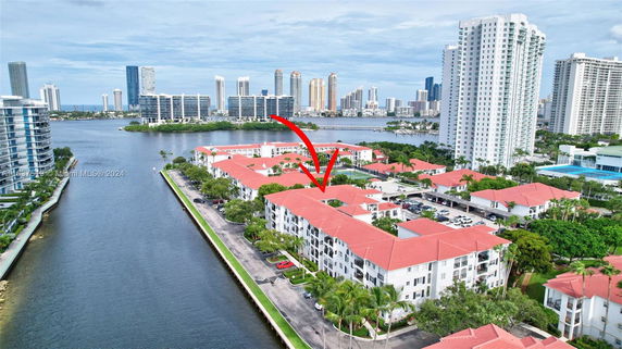 Aerial view of a waterfront residential area with multiple buildings and red roofs, surrounded by water and high-rise buildings in the distance.