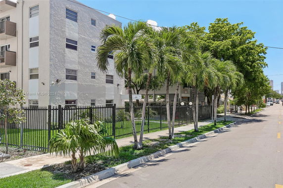 Front view of a multi-story apartment building with a black metal fence and palm trees.