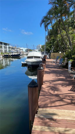 View of a boat docked along a waterside boardwalk with seating, featuring nearby buildings and clear sky.