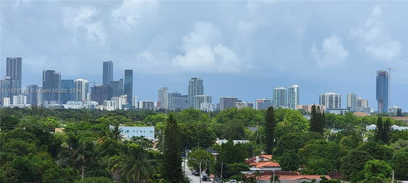 Cityscape view with tall buildings and greenery in the foreground under a cloudy sky.