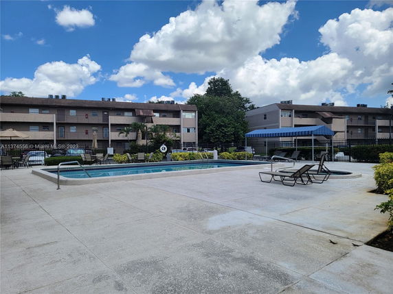 Outdoor swimming pool area with deck chairs and a shaded seating area near a multi-story residential building.