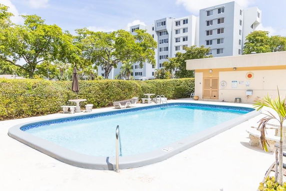 Outdoor swimming pool with lounge chairs and a surrounding hedge, with a multi-story building in the background.