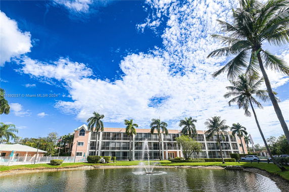 Front view of a multi-story residential building with palm trees and a pond with a fountain in the foreground.