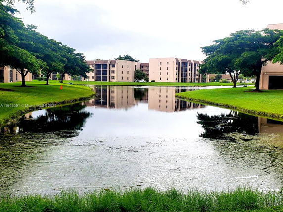 View of multiple apartment buildings surrounding a pond with grassy areas and trees.