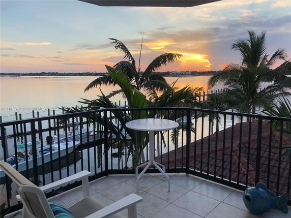 View from a balcony overlooking a calm body of water, with a sunset in the background and palm trees visible.