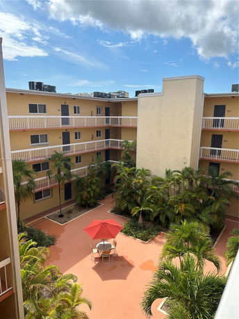 View of a multi-story building with balconies and a courtyard with palm trees and a table with an umbrella.