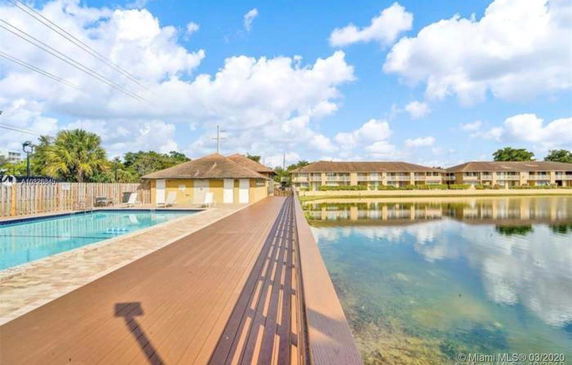 Outdoor swimming pool area with adjacent buildings and a pathway across a body of water.