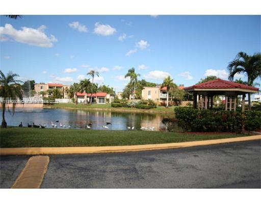 View of buildings with a pond and trees in the foreground under a blue sky.