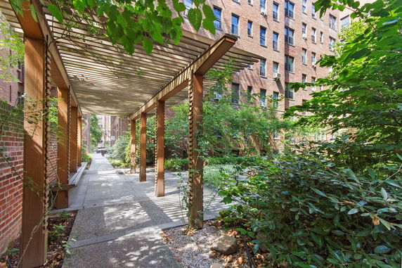 Pathway with a wooden pergola surrounded by greenery and adjacent brick buildings.