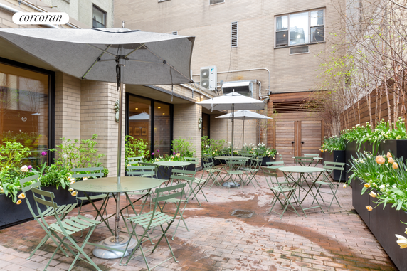 Outdoor patio area with tables, chairs, and umbrellas surrounded by greenery and potted plants.