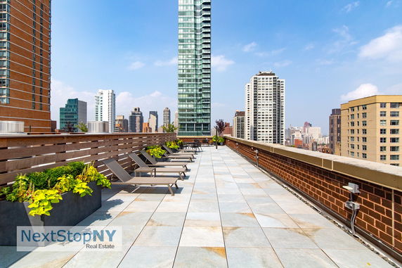 Rooftop terrace with lounge chairs and planters, overlooking city buildings.