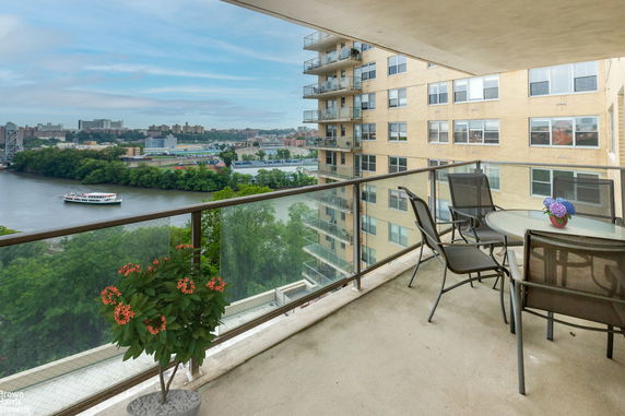 Balcony view from an apartment with a table and chairs, overlooking a river and a cityscape in the background.
