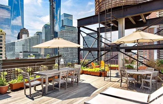 Rooftop area with wooden deck, dining tables, umbrellas, and a water tower, set against a city skyline.