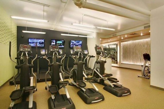 Indoor gym area with exercise machines and multiple screens.