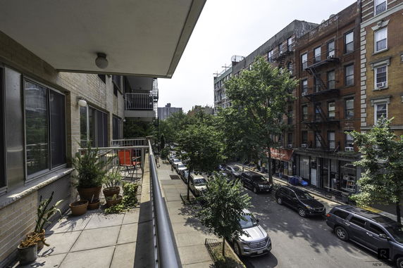 View from an apartment balcony overlooking a street with parked cars, trees, and surrounding buildings.