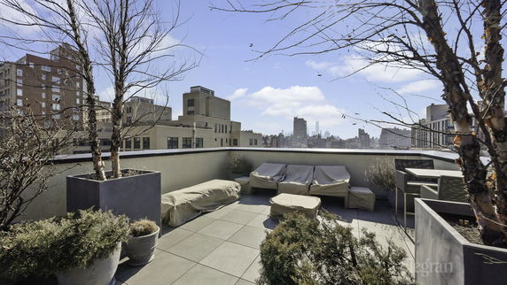 Rooftop area with patio furniture covered in protective covers and potted plants, surrounded by cityscape views.