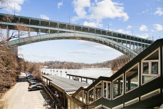 View of a bridge spanning over a river with a structure on the right and parked cars on the left.