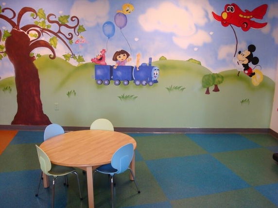 A small kids' play area with colorful chairs and a table set against a mural wall featuring cartoon characters.