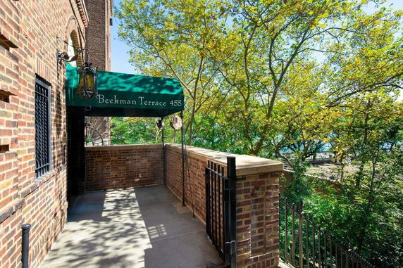 Entrance area of a brick building, with a green awning above the sidewalk, and trees in the background.