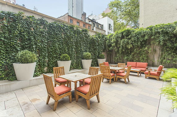 Outdoor seating area with wooden tables and chairs surrounded by tall greenery and potted plants.