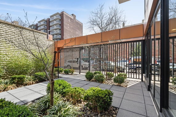 Outdoor area with tiled flooring and planters, bordered by a metal fence, adjacent to a building.