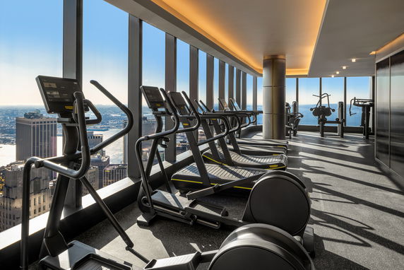Interior view of a gym with exercise equipment and large windows offering a cityscape view.