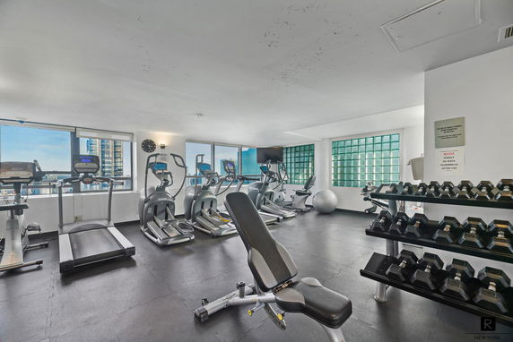 Interior view of a gym area with exercise equipment including treadmills, elliptical machines, a bench, and dumbbells.