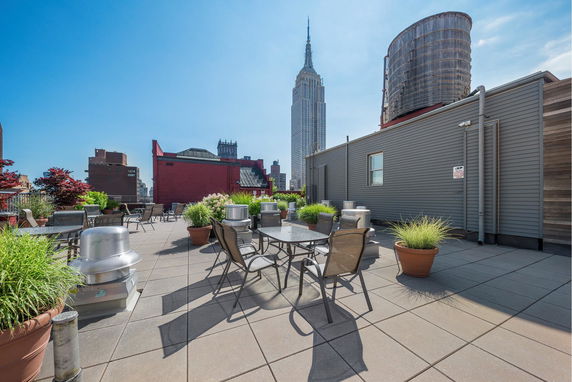 A rooftop area with tables, chairs, and potted plants, featuring views of nearby buildings.