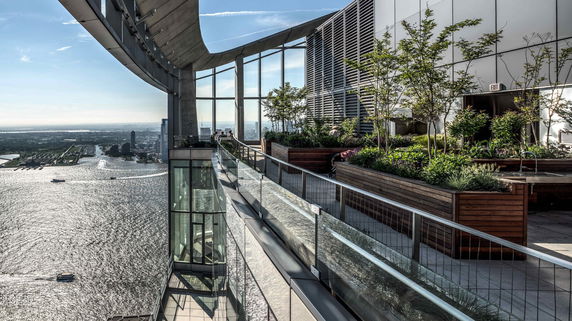 Rooftop area with plants and glass railings overlooking water and city.