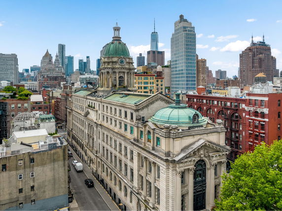 Aerial view of a large historical building with a green dome, surrounded by other urban buildings and skyscrapers.