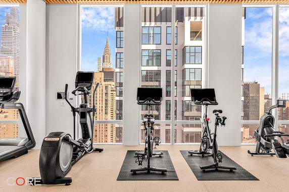 Indoor gym with exercise equipment and large windows showing a cityscape.