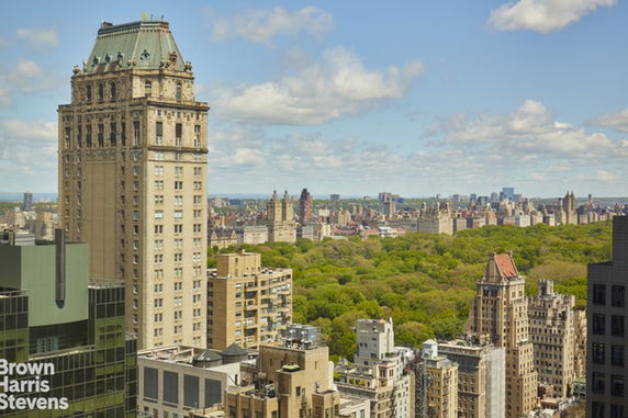 Aerial panoramic view of a cityscape featuring a tall historic building and a green park surrounded by other buildings.