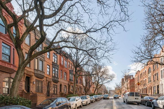 Street view of lined brownstone buildings with parked cars and bare trees.