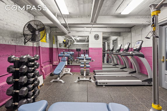Indoor gym area with dumbbells, benches, and treadmills against a pink and gray wall.