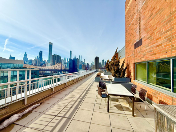 Rooftop area with tables and chairs on a building, offering a view of a city skyline and a bridge.