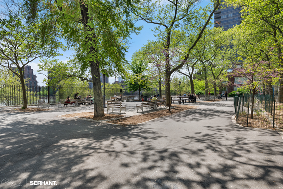 A city park area with benches, pathways, and trees, with buildings in the background.
